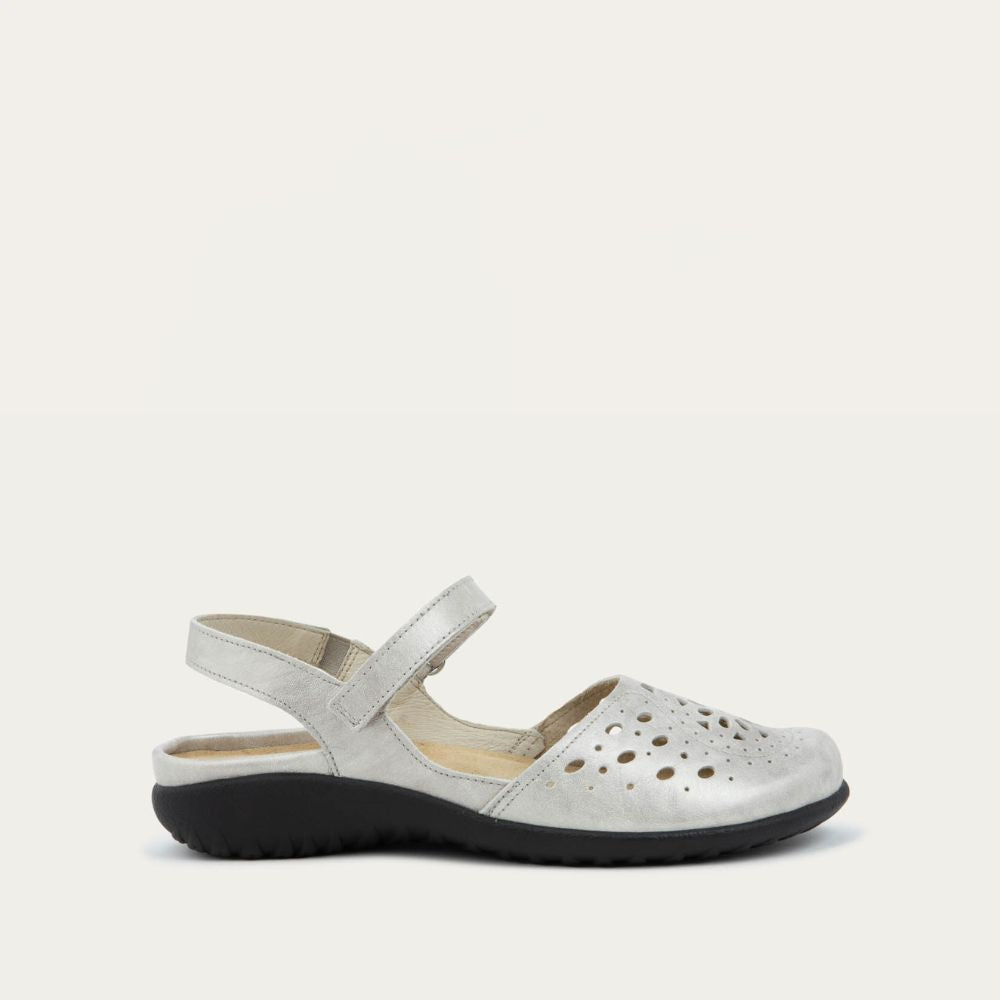 Profile view of a silver flat shoe with a perforated design, open heel, and adjustable ankle strap on a black sole