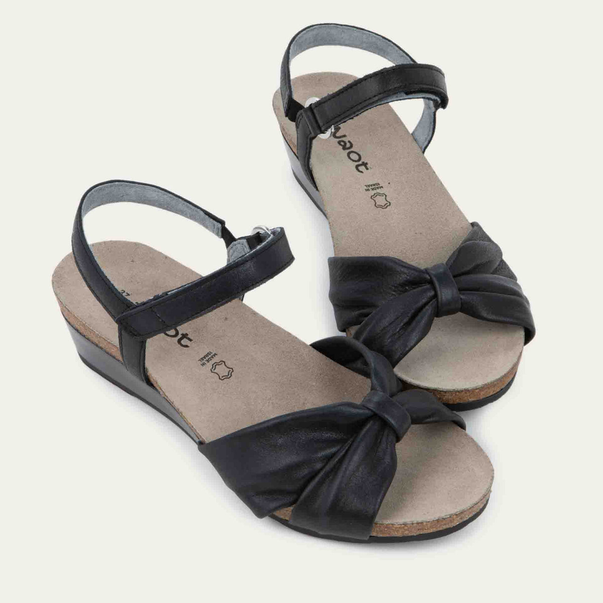 A pair of  black wedge sandals with an ankle strap and twisted front strap, featuring a cork footbed and wedge heel. 