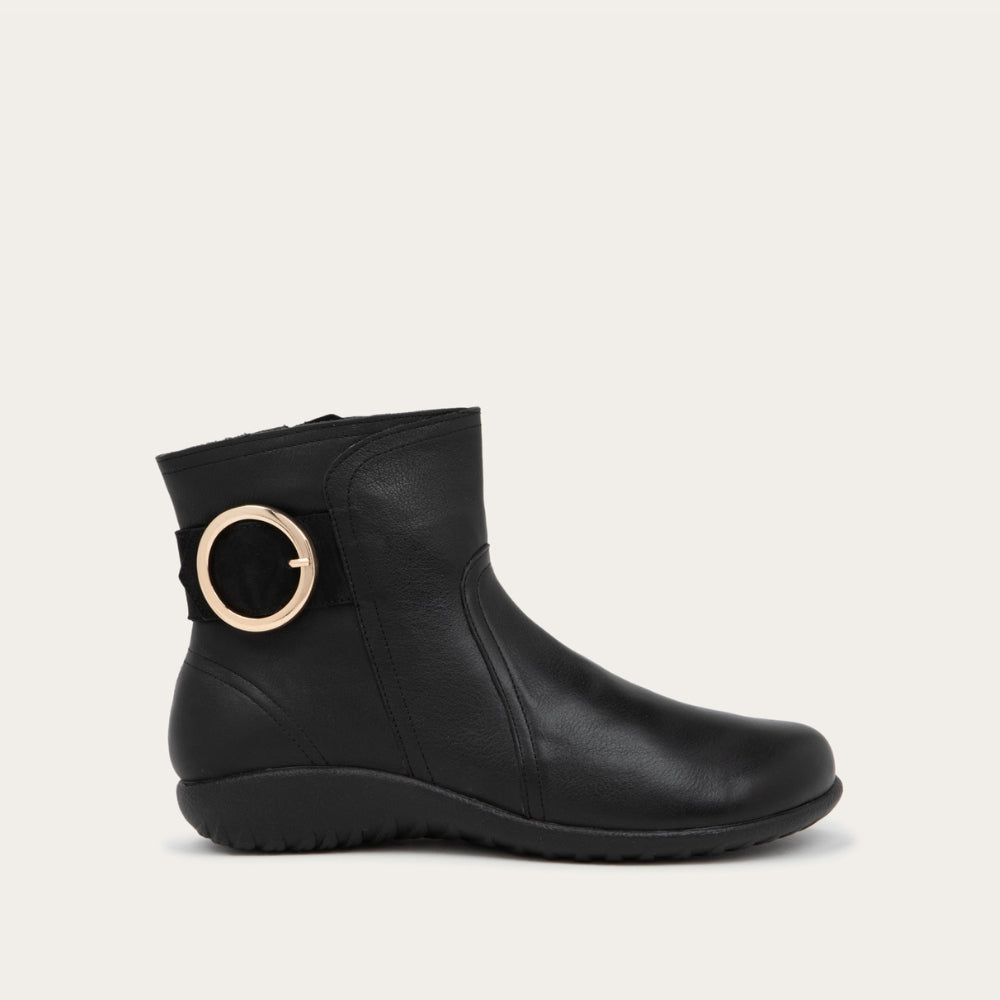 Profile view of a black boot featuring an accent strap with round buckle in the back of the shoe on a dark sole.
