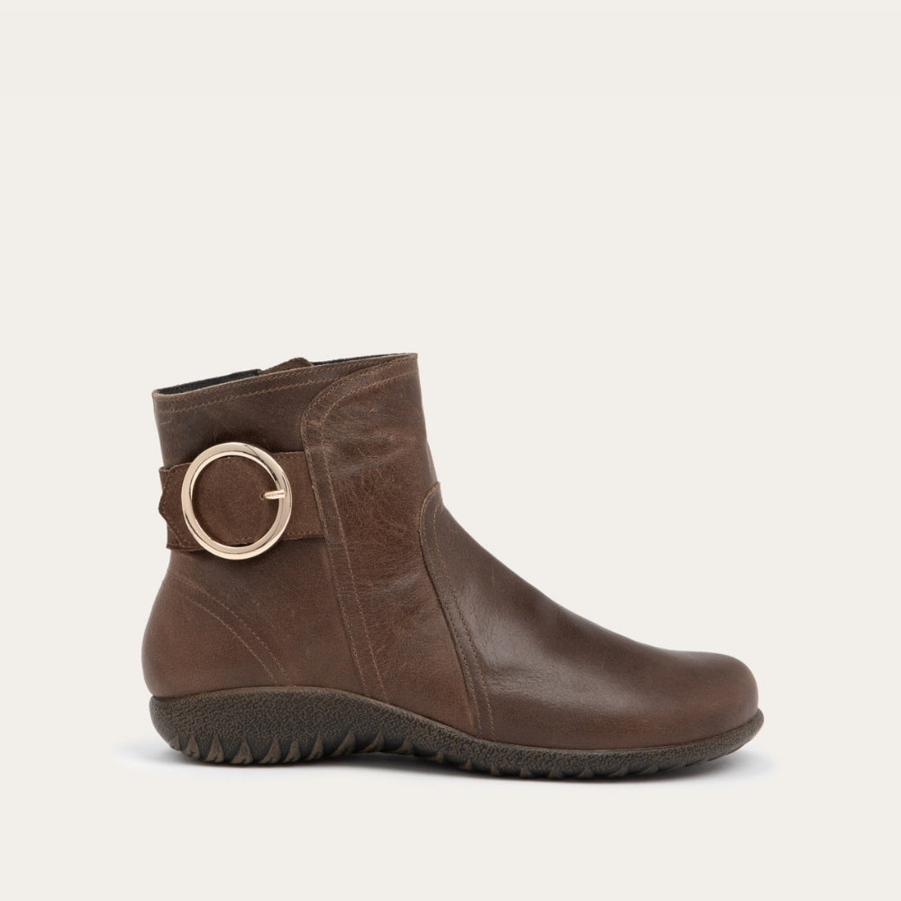 Profile view of a brown boot featuring an accent strap with round buckle in the back of the shoe on a dark sole.