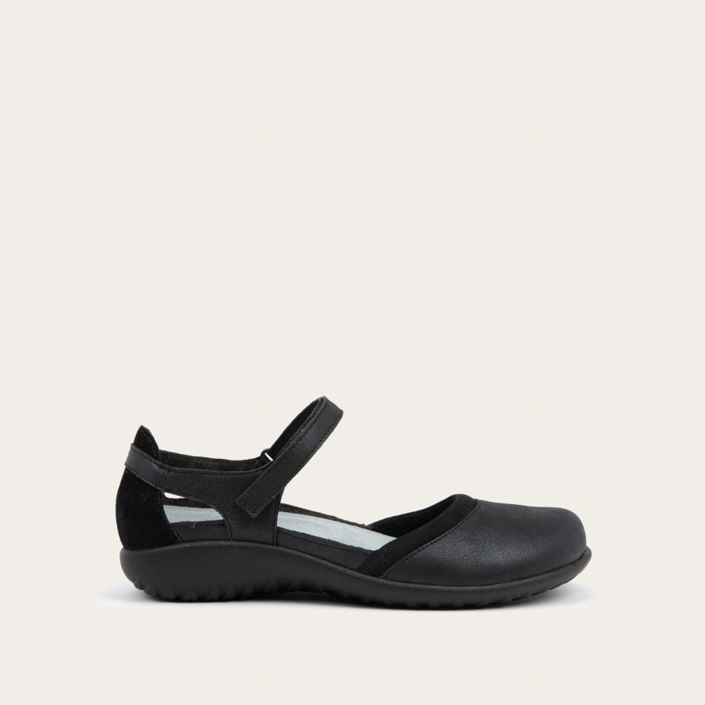 Profile view of a black Mary Jane featuring contrasting materials and cutouts by the heel on a dark sole.