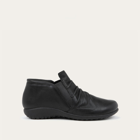 Side view of a black leather ankle boot with a textured sole and subtle ruching on the upper, set against a plain white background. The style is sleek and modern.